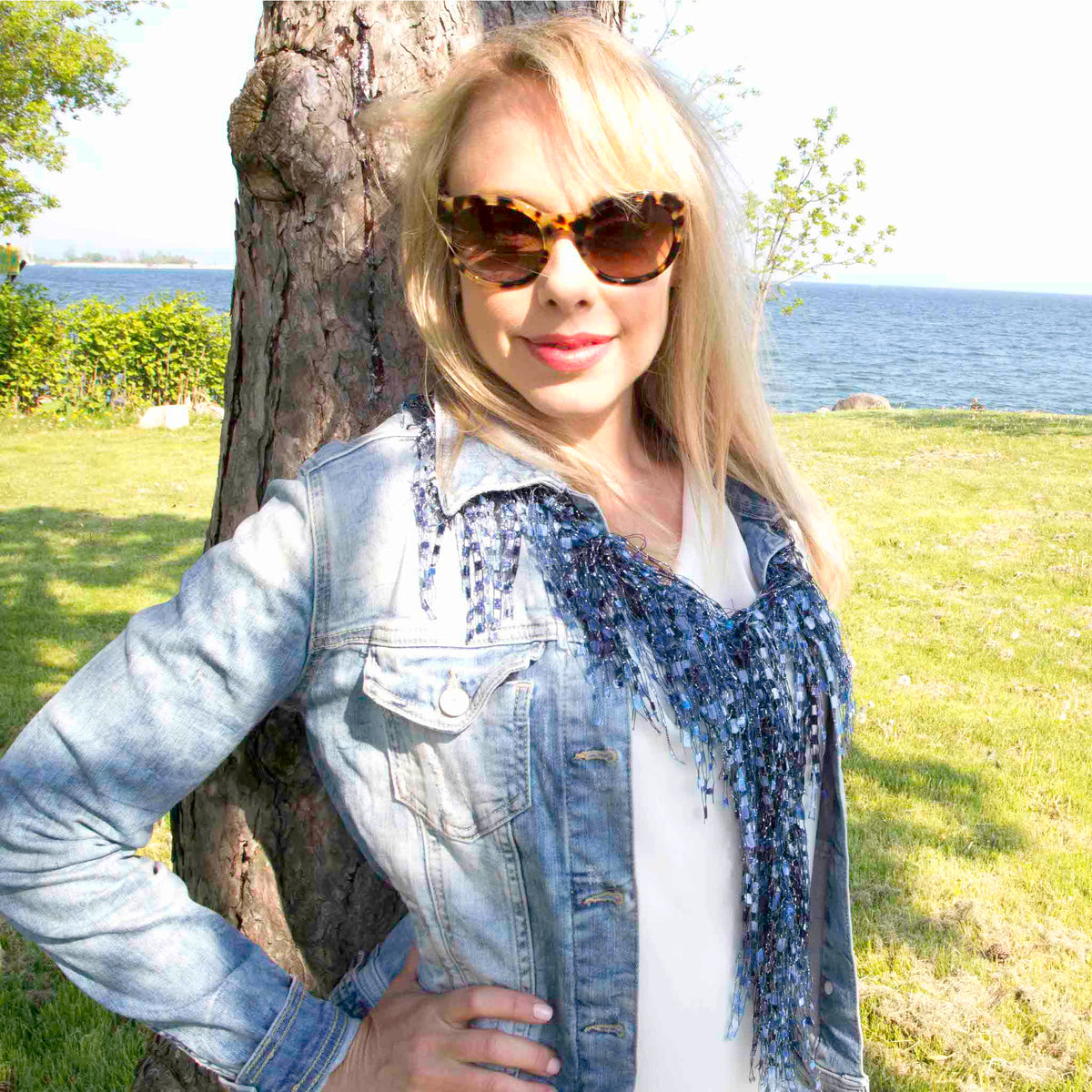Lady wearing a denim blue color Scarf Necklace worn behind the collar of denim jacket and white top. Lady is leaning against a tree in a park with water in the background. 